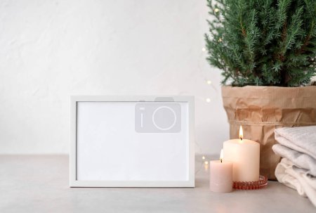 Photo for Blank white picture frame mockup on table, candles, juniper in pot, wool sweaters, white wall background. Cozy winter home interior decoration with Christmas lights, aesthetic holiday template. - Royalty Free Image