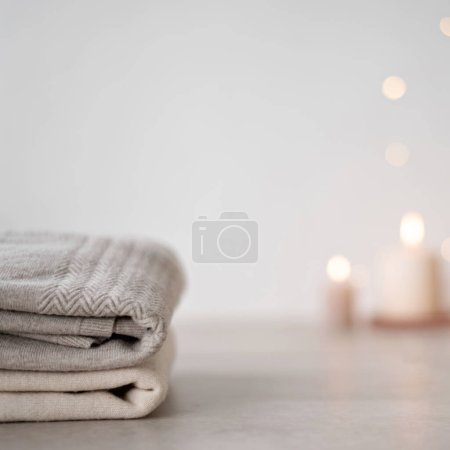 Foto de Suéteres de lana se apilan en la mesa, borrosa vela desenfocada luces de Navidad bokeh sobre fondo de pared blanco neutro. Mínima marca de negocio de invierno de vacaciones, plantilla de diseño blog. - Imagen libre de derechos