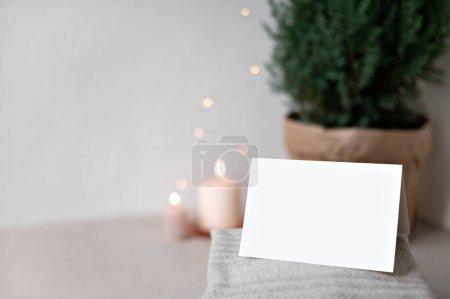 Foto de Tarjeta de papel en blanco maqueta, suéteres en la mesa, velas desenfocadas borrosas y luces de guirnalda, enebro en maceta en el fondo. Espacio de copia de pared beige neutro vacío. Tarjeta postal de Navidad o plantilla de invitación - Imagen libre de derechos