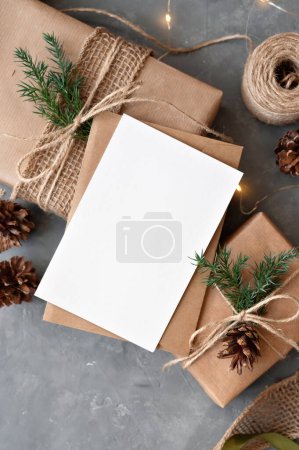 Foto de Tarjeta de papel en blanco maqueta, sobre, cajas de regalo decoradas con papel hecho a mano, ramitas de pino y conos. Celebración de Navidad sostenible, hacer regalos, felicitación postal, plantilla de invitación. - Imagen libre de derechos
