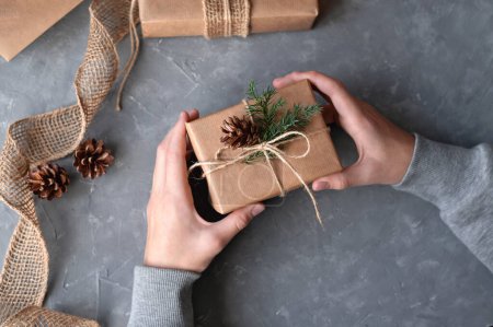 Foto de Manos sosteniendo caja de regalo de Navidad decorada con conos de coníferas y ramitas, envuelta en papel hecho a mano, sobre fondo de mesa gris. - Imagen libre de derechos