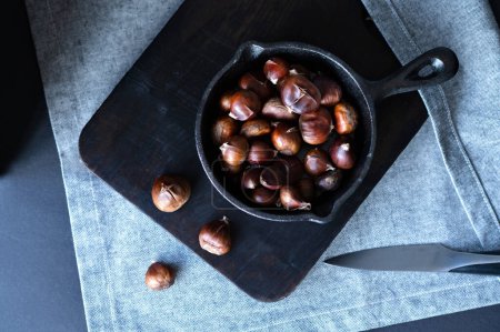 Photo for Hot roasted chestnuts in pan, on black wooden board, on kitchen table. Traditional Christmas food, home cooking. - Royalty Free Image