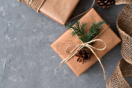 Téléchargez les photos : Cadeaux de Noël esthétiques sur fond de table gris. Boîtes cadeaux marron décorées de papier crafter, de brindilles de conifères et de pins. Concept durable de célébration écologique des vacances d'hiver. - en image libre de droit