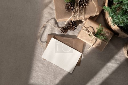 Téléchargez les photos : Carte papier vierge, maquette d'enveloppe, coffrets cadeaux de Noël fabriqués sur nappe beige neutre en lin avec ombres naturelles, carte postale de Noël esthétique, modèle d'invitation, style champêtre nordique - en image libre de droit