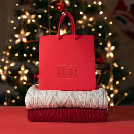 Foto de Suéteres de punto rojo y blanco se apilan en la mesa roja, mockup bolsa de compras brillante con espacio de copia vacío, árbol de Navidad borrosa con luces en el fondo. Compras de vacaciones de invierno, ventas, concepto de regalos. - Imagen libre de derechos