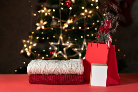 Foto de Maqueta de tarjeta de regalo de vacaciones de invierno para compras de venta de Navidad, suéteres de punto, bolsa de compras roja y tarjeta de papel en blanco en la mesa roja, árbol de Navidad borrosa brillante en el fondo, plantilla de marca. - Imagen libre de derechos