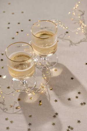 Photo for Aesthetic New Year festive background, glasses with sparkling wine on neutral beige linen tablecloth with gold confetti and natural sunlight shadows, soft selective focus, lifestyle. - Royalty Free Image