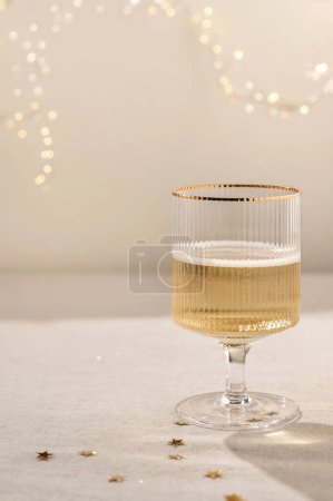 Téléchargez les photos : Verre à vin avec alcool de vin mousseux boisson sur nappe en lin beige avec ombres naturelles et confettis d'or, guirlande floue lumières sur le fond. Esthétique Nouvel An, mariage, fond d'anniversaire. - en image libre de droit