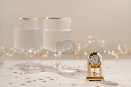 Photo for New Year's eve celebration concept, golden clock with twelve o'clock time, midnight, on beige table with gold star confetti, blurred wineglasses with sparkling wine and garland lights on background. - Royalty Free Image