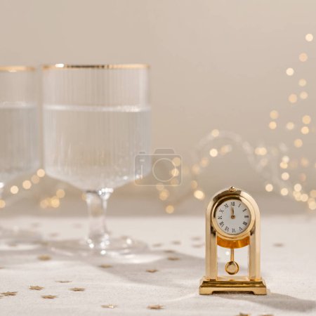 Foto de Tiempo para el concepto de celebración de Año Nuevo, reloj de época de oro que muestra las doce, gafas de vino con vino espumoso, luces de guirnalda borrosa en el fondo. Fondo festivo estético neutro. - Imagen libre de derechos