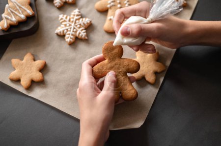 Photo for Christmas gingerbread cookie decoration. Hands holding gingerbread man cookie and piping bag with icing, making cookie decor. Winter holiday traditional family cooking. Selective focus, lifestyle. - Royalty Free Image