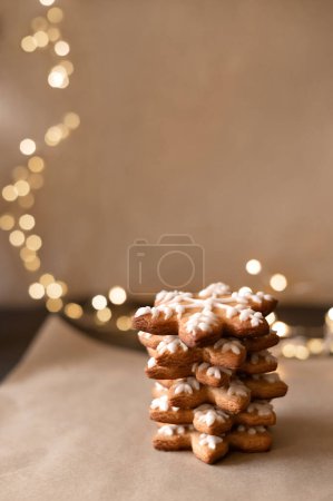 Téléchargez les photos : Pain d'épice en forme de flocon de neige pile de biscuits sur parchemin beige, lumières de guirlande floues brillantes sur le fond. Dessert doux maison traditionnel de Noël, foyer sélectif doux, style de vie. - en image libre de droit