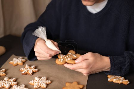 Téléchargez les photos : Garçon en pull foncé décorant biscuit homme pain d'épice avec glaçage au sucre, tenant dans les mains sac de tuyauterie et biscuit. - en image libre de droit