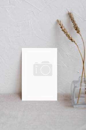 Foto de Plantilla de diseño de póster interior minimalista o marco de imagen, maqueta de tarjeta de papel en blanco sobre mesa beige neutra, jarrón con hierba seca del prado, fondo de pared texturizado de yeso blanco vacío. - Imagen libre de derechos