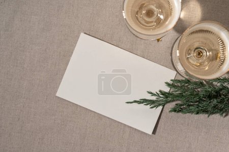 Foto de Año Nuevo, invitación al evento de vacaciones de invierno o plantilla de tarjeta postal de felicitación, maqueta de tarjeta de papel en blanco sobre fondo de mesa de lino beige neutro, gafas de vino con vino espumoso y rama de abeto. - Imagen libre de derechos