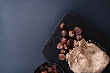 Foto de Castañas asadas comestibles en saco de yute esparcidas sobre tabla de madera marrón, sobre fondo gris oscuro con espacio para copiar. Vista superior, plano. - Imagen libre de derechos