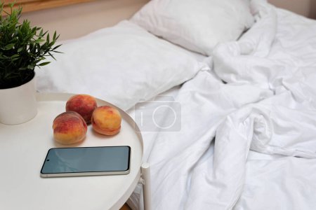 Photo for Empty messy unmade bed with white pillows and blanket, bedside table with peaches, mobile phone and plant in pot, lifestyle home bedroom interior. - Royalty Free Image