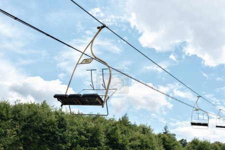 Téléchargez les photos : Téléphérique avec sièges de téléphérique vides dans le ciel bleu, au-dessus de forêt de pins. Voyage en montagne en été. - en image libre de droit