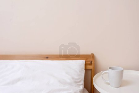 Téléchargez les photos : Lifestyle minimaliste chambre à coucher détails intérieurs, lit avec oreiller blanc. tasse de café sur la table de chevet, fond de mur neutre pêche vide. - en image libre de droit