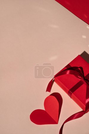 Foto de Caja de regalo roja y tarjeta de San Valentín en forma de corazón de papel sobre fondo beige melocotón con luz brillante y sombras duras. Concepto de celebración mínima de San Valentín, plantilla de marca comercial. - Imagen libre de derechos