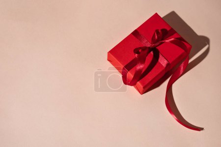 Foto de Caja de regalo roja con cinta sobre fondo de melocotón pastel en luz brillante con sombras duras, celebración mínima de vacaciones de lujo, saludo presente telón de fondo con espacio para copiar. - Imagen libre de derechos