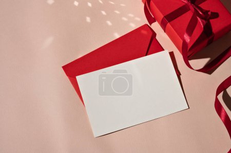 Photo for Luxury feminine festive greeting postcard template, blank paper card mockup, red envelope and gift box on pastel pink background with bright light and shadows. Holiday celebration backdrop. - Royalty Free Image