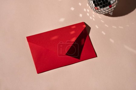 Téléchargez les photos : Enveloppe en papier rouge et boule miroir étincelante sur fond beige avec reflets lumineux et ombres, carte de vœux minimaliste esthétique, invitation, concept de lettre. - en image libre de droit