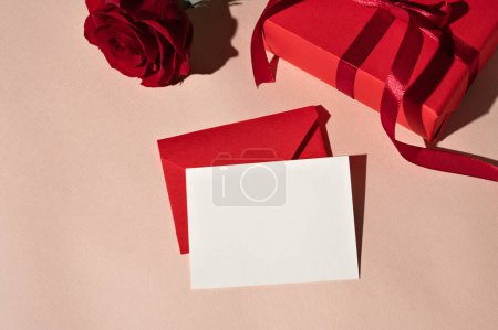 Téléchargez les photos : Modèle de carte de papier vierge, fleur de rose rouge, boîte cadeau, enveloppe sur fond rose pastel, carte de vœux de la Saint-Valentin ou de la fête des mères, modèle d'invitation à un événement de mariage. - en image libre de droit