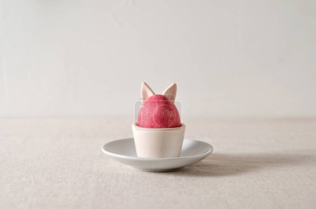 Foto de Concepto minimalista estético de vacaciones de Pascua, huevo de color rosa en taza con orejas de conejo en plato, sobre fondo de lino beige neutro, enfoque selectivo suave, luz natural del sol y sombras. - Imagen libre de derechos