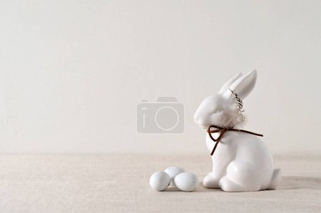 Foto de Fondo minimalista estético de vacaciones de Pascua, decoración de conejito de porcelana blanca y pequeños huevos de caramelo en mantel de lino beige neutro claro, luz solar natural suave, enfoque selectivo. - Imagen libre de derechos