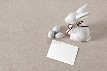 Foto de Minimalista tarjeta postal de felicitación de vacaciones de Pascua, invitación o plantilla de tarjeta de visita, maqueta de tarjeta de papel en blanco, figurilla de conejo de cerámica y pequeños huevos sobre fondo de mantel de lino taupe neutro. - Imagen libre de derechos