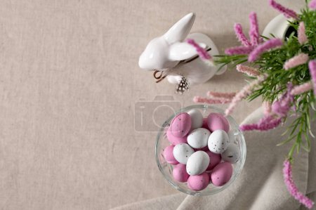 Téléchargez les photos : Décor de vacances de Pâques et bonbons, petits bonbons roses et blancs en forme d'oeuf dans un vase, figurine de lapin, fleurs floues sur fond de nappe en lin beige neutre, vue sur le dessus. - en image libre de droit