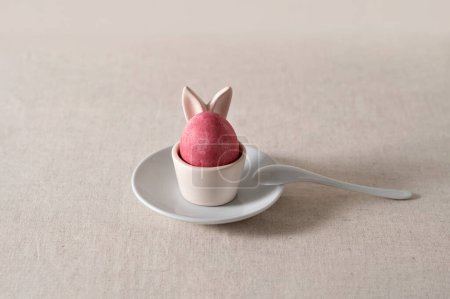 Foto de Decoración de comida de Pascua, huevo de color rosa en taza de huevo con orejas de conejo en platillo con cuchara, ajuste de mesa minimalista de Pascua, enfoque selectivo suave. - Imagen libre de derechos
