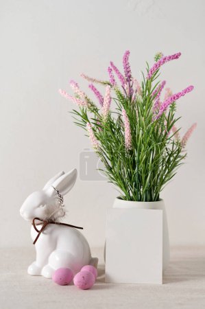 Photo for Easter holiday greeting postcard or invitation template, blank paper card mockup, flowers in vase, rabbit figurine, pink candy eggs on neutral beige background, soft natural light. - Royalty Free Image
