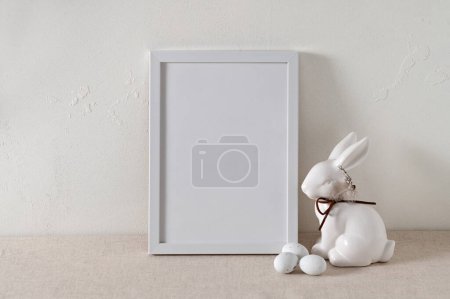 Foto de Plantilla de cartel de vacaciones de Pascua blanca minimalista, maqueta de marco de imagen en blanco, figura de conejo y pequeños huevos de caramelo sobre fondo de mantel de lino beige neutro, pared de hormigón vacío texturizado. - Imagen libre de derechos
