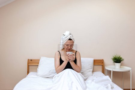 Foto de Mujer serena adulta con toalla de turbante en la cabeza y taza de café en las manos, sentado en la cama en el dormitorio beige melocotón, rutina de la mañana estilo de vida saludable, autocuidado, emoción positiva. - Imagen libre de derechos