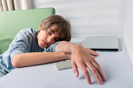 Photo for Young teenage handsome boy sleeping, laying head on hands on tabletop, with mobile phone and laptop nearby. Gadget detox, rest and relaxation, break from electronic devices. Lifestyle home interior. - Royalty Free Image