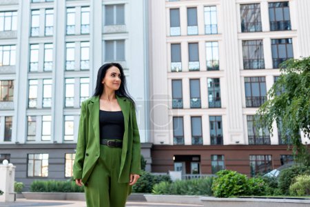 Foto de Hermosa mujer morena adulta confiada vistiendo traje de moda verde, chaqueta y pantalones, caminando por la calle urbana y sonriendo, mujer de negocios. - Imagen libre de derechos