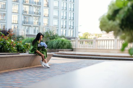 Téléchargez les photos : Femme brune adulte en costume de mode verte assise sur un banc sur la place de la ville urbaine, avec bouquet de fleurs d'hortensia, tenant la main sur les pieds. - en image libre de droit