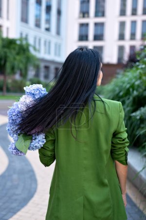 Téléchargez les photos : Femme brune adulte élégante portant une veste verte avec bouquet de fleurs d'hortensia bleues marchant dans la rue de la ville, avec les cheveux en mouvement, vue de dos, streetstyle, foyer sélectif doux. - en image libre de droit