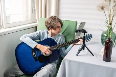 Téléchargez les photos : Adolescent garçon blogueur jouer de la guitare et enregistrement vidéo sur smartphone, streaming en ligne pour les médias sociaux blog ou webinaire leçon. - en image libre de droit