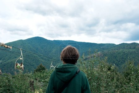 Téléchargez les photos : Homme regardant le paysage de montagne avec des pins, ciel nuageux et téléphérique, debout au sommet de la montagne, vue de dos. Montagnes des Carpates, Ukraine. - en image libre de droit