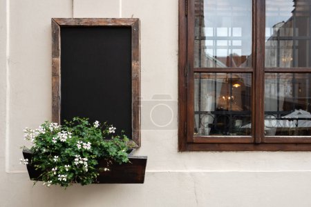 Photo for Blank chalkboard with a wooden frame, next to window, adorned by flower box with blooming white flowers. Authentic vintage cafe building facade. - Royalty Free Image