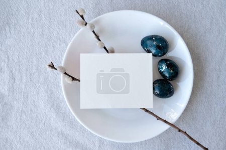 Foto de Minimal aesthetic Easter holiday invitation design template, blank paper card mockup, pussy willow, blue dyed eggs on white plate, on neutral linen tablecloth background. - Imagen libre de derechos