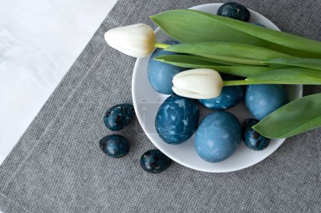 Téléchargez les photos : Assiette avec oeufs de Pâques teints en bleu et fleurs de tulipe sur fond de serviette en lin gris, composition des vacances de Pâques, style scandinave. - en image libre de droit