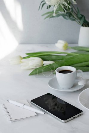 Photo for Elegant home office workspace, mobile phone, notebook, pen, cup of coffee and flowers on white table with natural sunlight, business morning concept. - Royalty Free Image