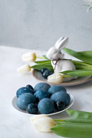 Photo for Blue colored Easter Eggs, bunny figurine, tulip flowers on neutral white table background with soft natural light, aesthetic lifestyle Easter holiday home decoration in rustic style, selective focus. - Royalty Free Image