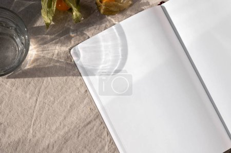 Photo for Blank paper texture, closeup of open empty notebook sheet mockup, glass with water, fruits on beige linen table cloth background with natural sunlight shadows, lifestyle business branding template. - Royalty Free Image