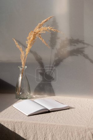 Photo for Aesthetic boho summer still life, home interior decoration, open book, vase with dried grass on table with sand beige linen textured tablecloth, harsh natural sunlight shadows. - Royalty Free Image