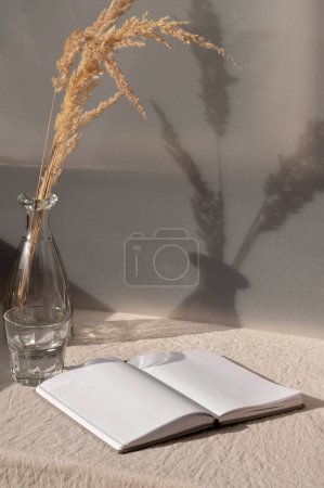 Foto de .Libro abierto y jarrón con hierba seca en la mesa con mantel de lino beige, boho boho verano naturaleza muerta, interior estético hogar sostenible en colores terrosos. - Imagen libre de derechos